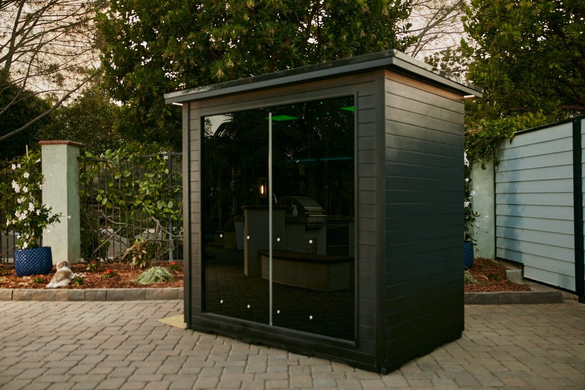 Black Sun Home Luminar 5 cabin with a flat overhanging roof and a two-panel tinted glass front, on a paver patio in a garden at dusk