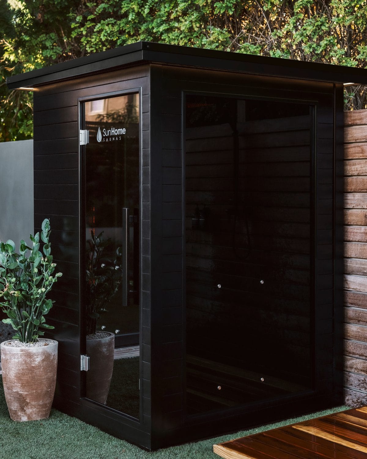 Black Sun Home Luminar 2 cabin with a flat overhanging roof, a glass door with the Sun Home logo and one large tinted glass side, on artificial turf beside a wood fence and a potted plant
