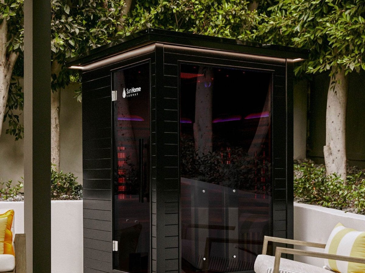 Black Sun Home Luminar 2 cabin with a flat overhanging roof, a glass door with the Sun Home logo and a tinted glass side, on a landscaped patio beside an outdoor chair