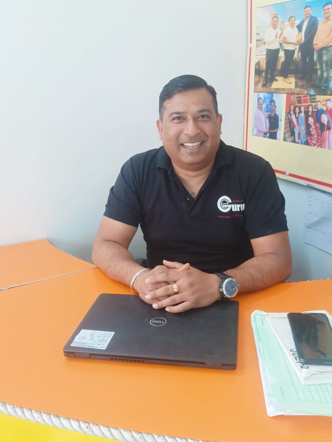 Circular studio-style portrait of a middle-aged South Asian man wearing a brown leather jacket over a black high-neck shirt, facing forward with a neutral expression against a plain light background.