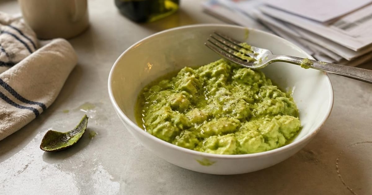 A slightly chipped ceramic bowl featuring mashed avocado with a light pinch of sea salt and a drizzle of olive oil, sitting next to a scattered pile of opened mail on a kitchen island.