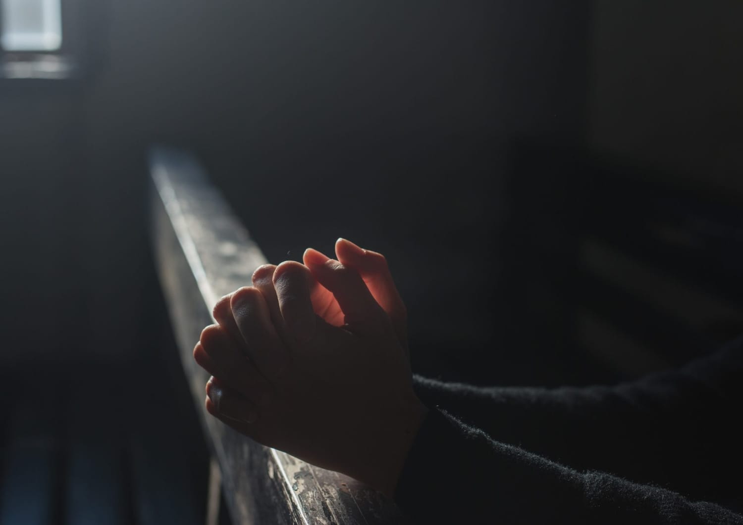 Hands folded in prayer on a wooden pew in low light