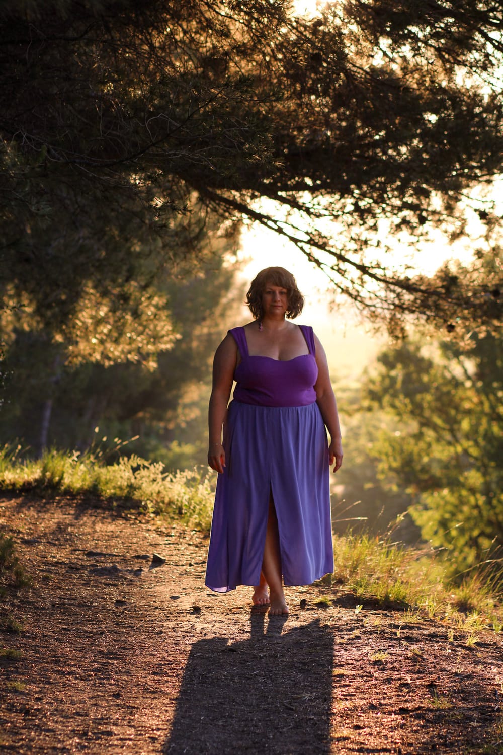 Photo de Céline Captin vêtue de violet avec une longue jupe fendue. Elle marche à contre jour dans un chemin boisé au soleil levant. Cette photo inspire la sérénité, le clame, la respiration, le bien-être, la pleine présence, la tranquilité et l'accomplissement.