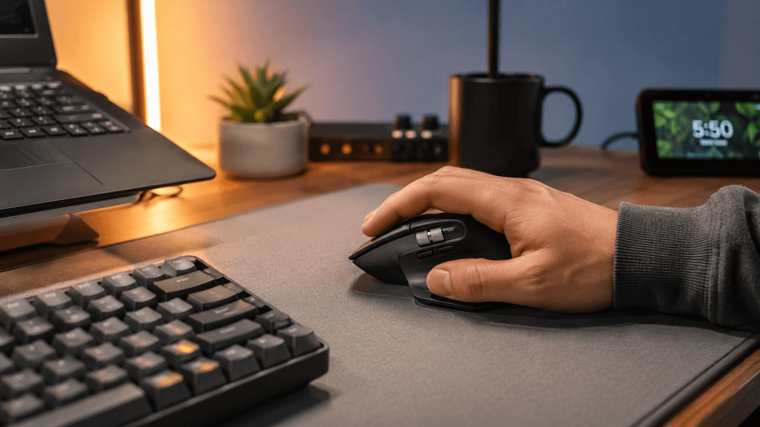 Close-up of a hand using an ergonomic wireless productivity mouse beside a compact mechanical keyboard on a minimalist desk mat with warm ambient lighting in a modern home office setup.