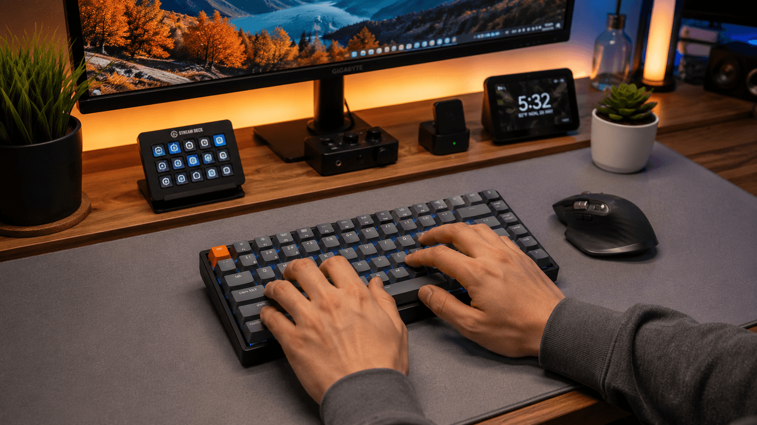 Hands typing on a compact wireless mechanical keyboard beside an ergonomic productivity mouse on a minimalist desk mat, with warm ambient lighting, modern monitor setup, and cozy work-from-home desk aesthetic.