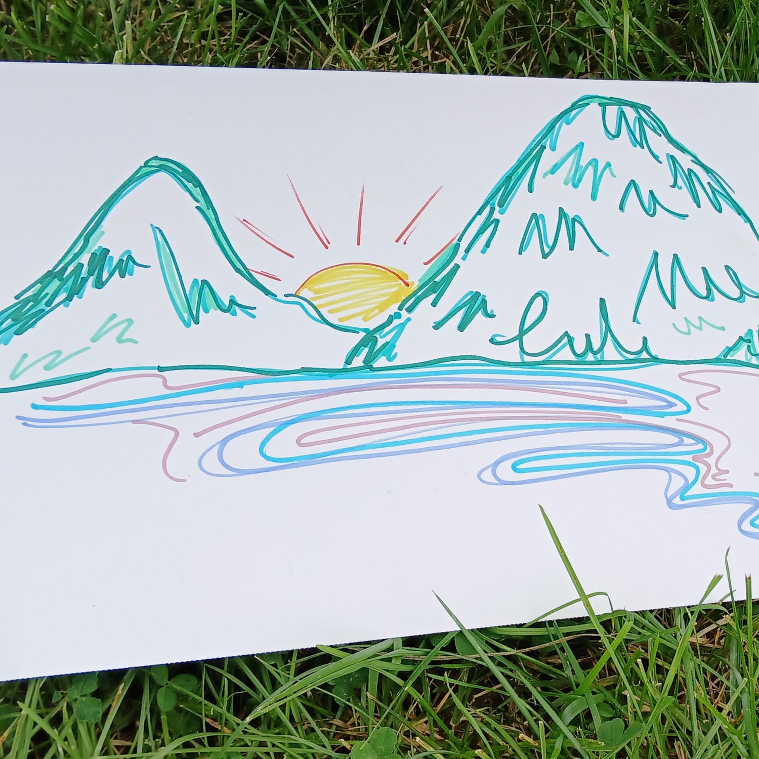 Marker drawing by Elissa: a sun rising between two green mountains over water, lying on the grass