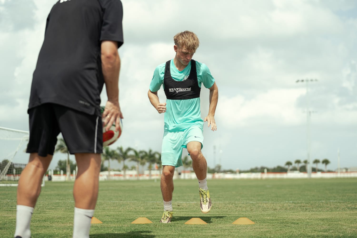 Youth player running an agility drill through orange cones while his coach waits with the ball