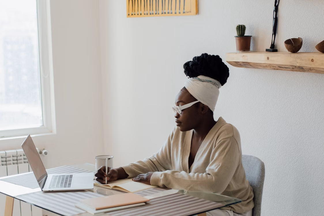 Une femme prend des notes à côté de son ordinateur dans un intérieur lumineux.