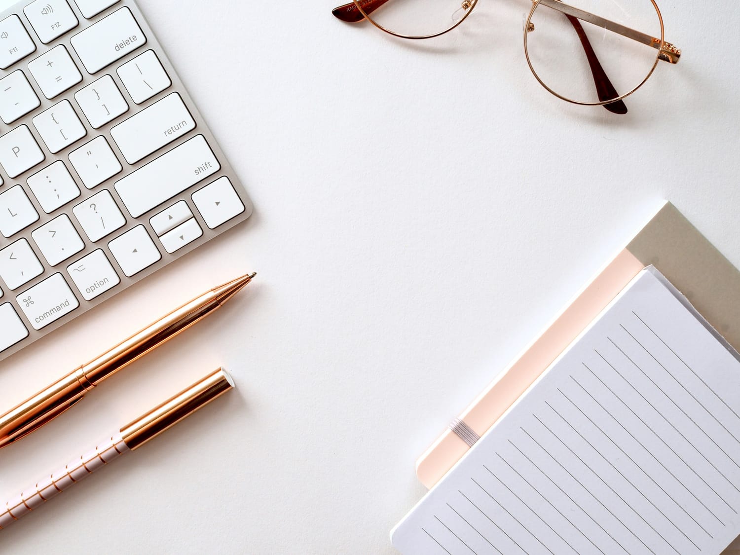A desk with a keyboard and rose gold writing utensils on the left, glasses on top and notepads on the bottom right.