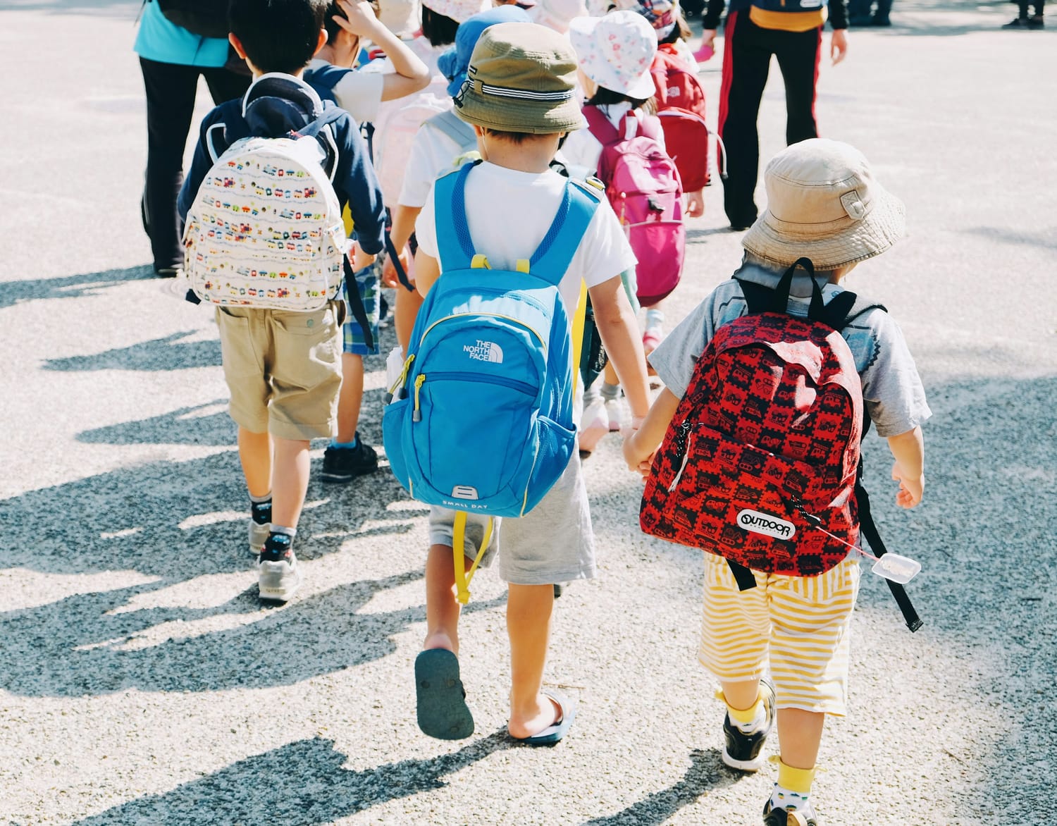 Group of young children in a line walking away from camera. They are wearing tan shorts and two are wearing tan hats. There is a cream, blue and red backpack on the kids you see.