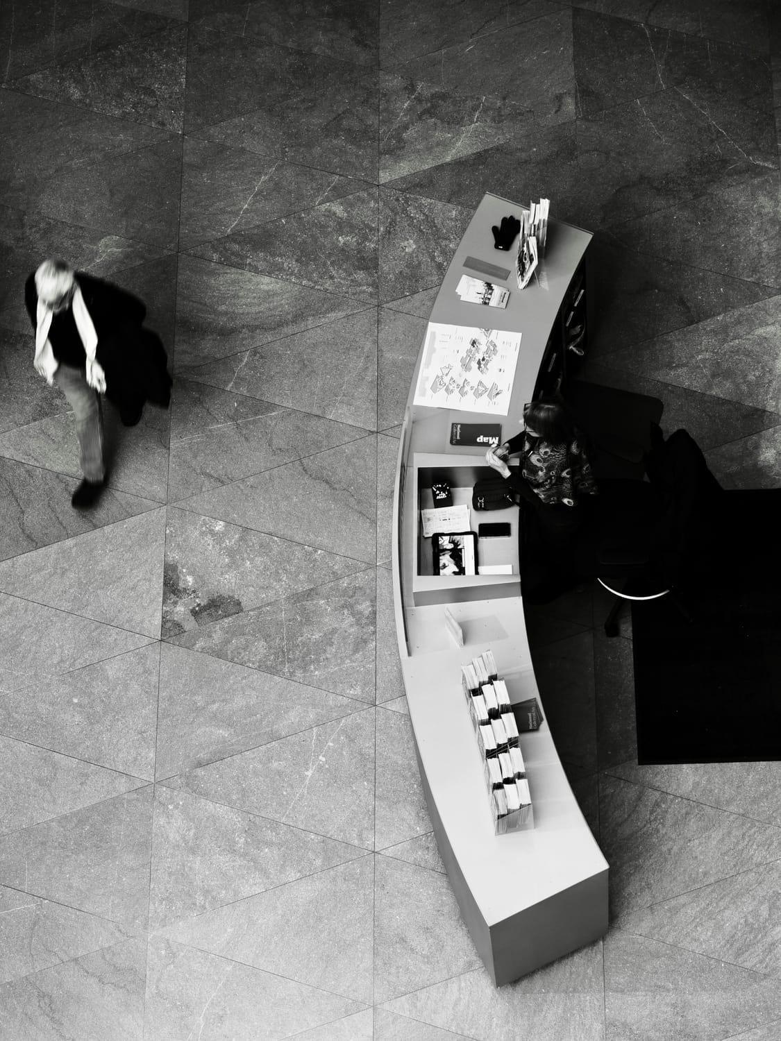 A grayscale photo overview of a person walking on the left. An arch shaped desk with brochures, other paper material s, and a person at the desk.