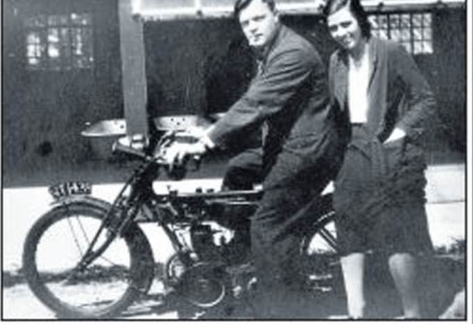 Emily Kathleen Renshaw smiles while leaning leans against a h=genytlemen sat on a motorbike wearing dark overalls with a cigerette hanging from his lips.