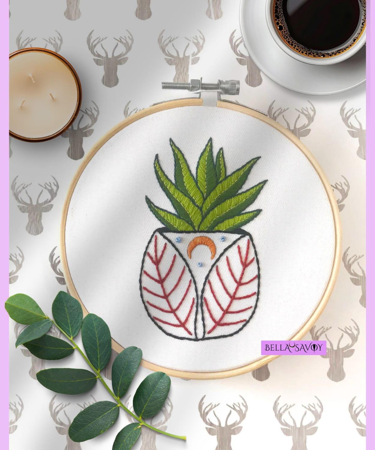 minimalist flatlay with a potted succulent embroidery on a hoop set against a white background with stag silhouette pattern. Surrounded by a cup of black coffee, a candle, and stem with leaves. Soft cast shadow