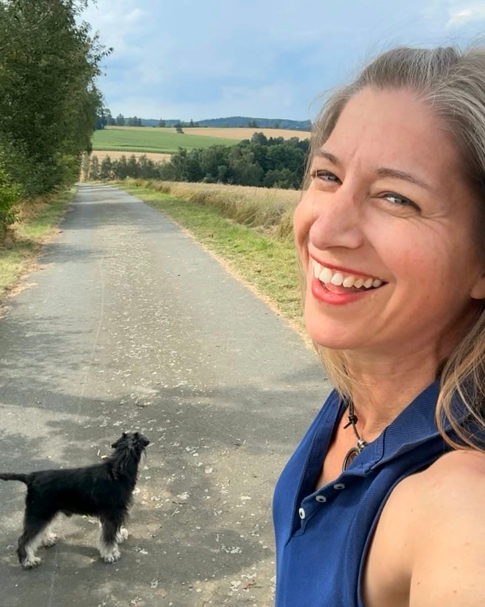 Kristin Voigt lacht in die Kamera, hinter ihr ein Feldweg und ihr Zwergschnauzer Pino