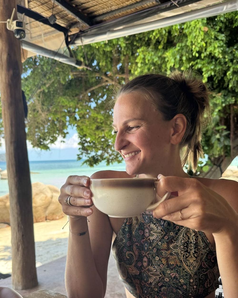 Nadia lächelt mit einer Tasse in der Hand am Strand