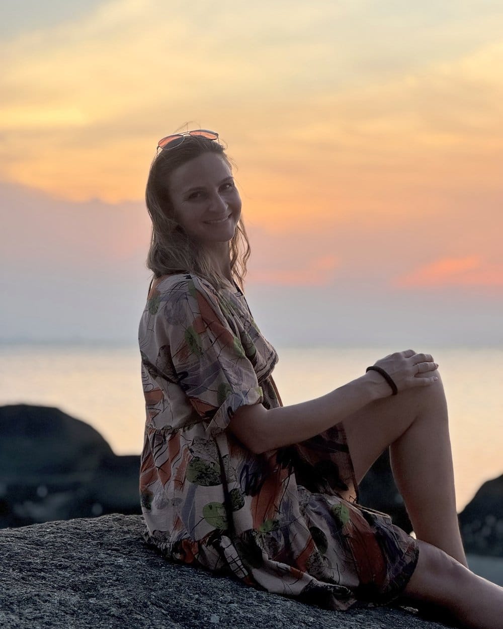 Nadia sitzt lächelnd auf einem Felsen am Meer im Sonnenuntergang