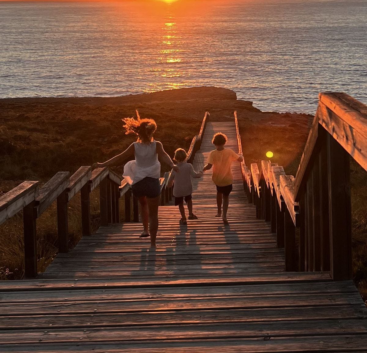 Eine Mutter läuft mit zwei Kindern Hand in Hand eine Holztreppe hinunter zum Meer, im Gegenlicht der Abendsonne.