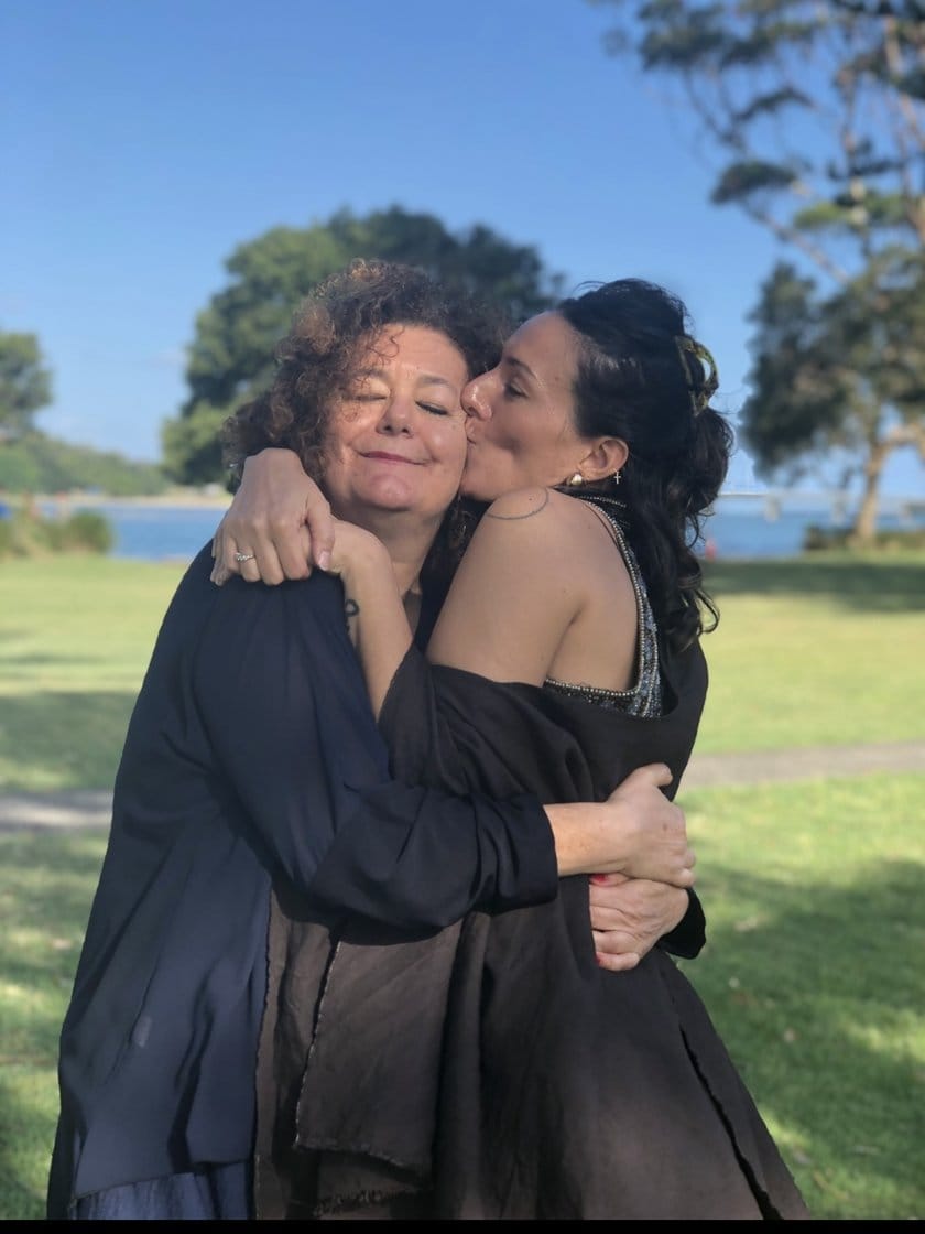 Sabrina Mascanzoni abbracciata dalla figlia Aurora, che le dà un bacio sulla guancia, in un parco davanti al mare