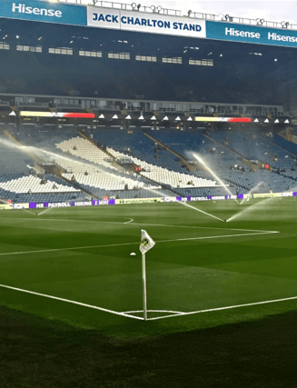 A Leeds United corner flag beside the pitch at Elland Road, with sprinklers watering the pitch and stadium seating behind.