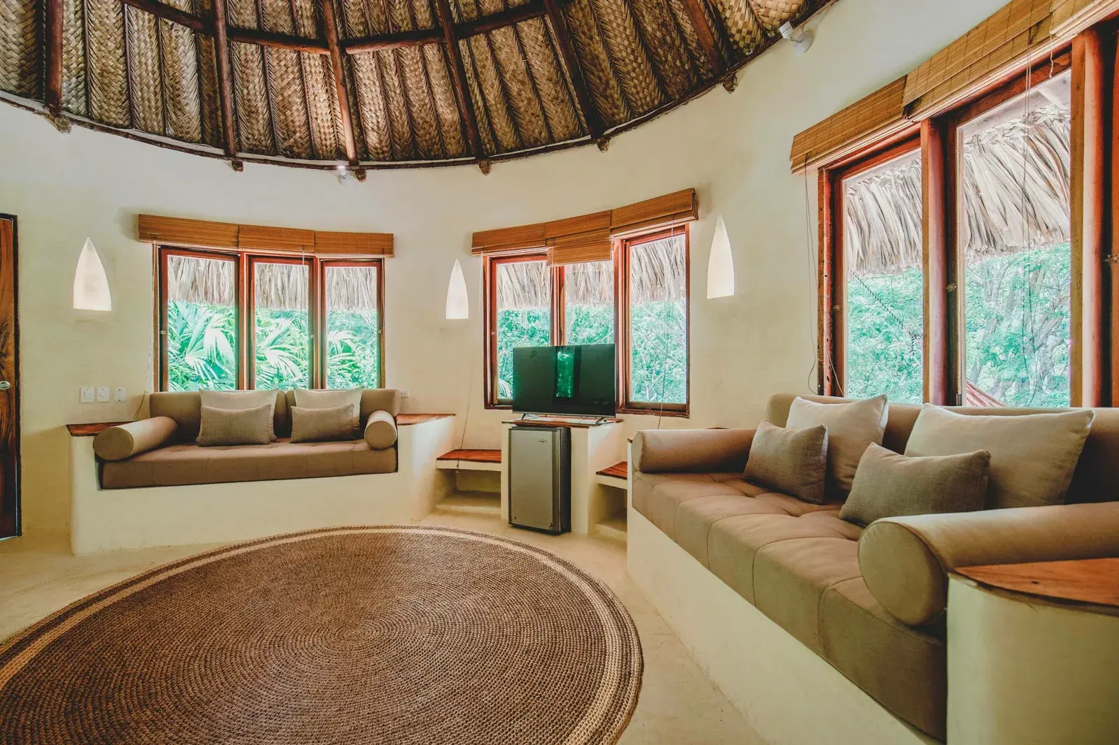 Sala de una cabaña con techo de palma y ventanas a la selva