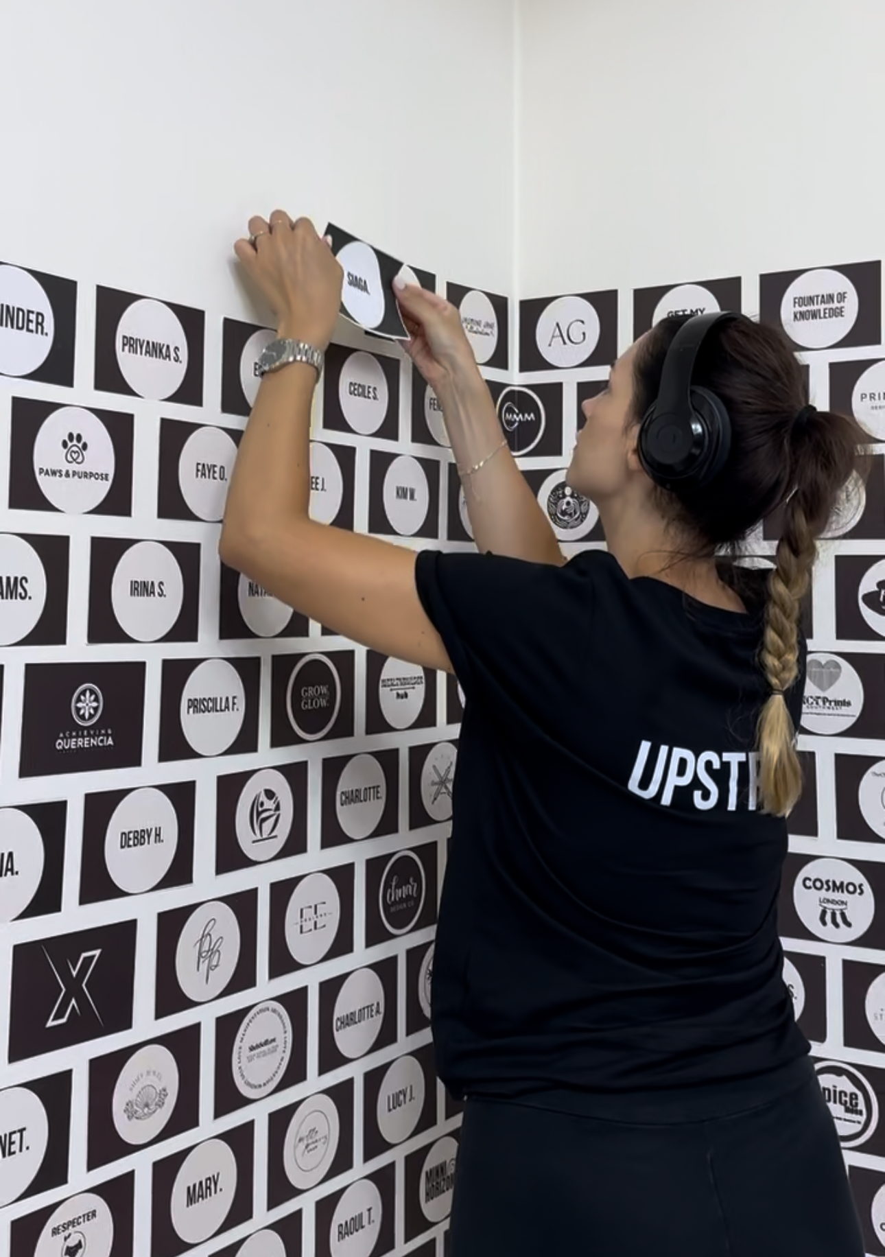 Anne wearing Upstrider t-shirt placing a member’s logo tile on a branded wall filled with logos from other Upstrider 30K+ Plan™ participants.