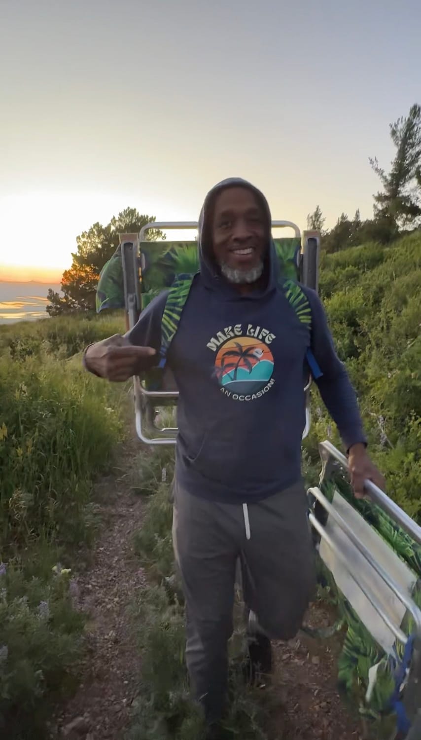 man smiling, pointing to his sweatshirt that shows a make life occasion logo walking with two camping charis with a sunset behind him