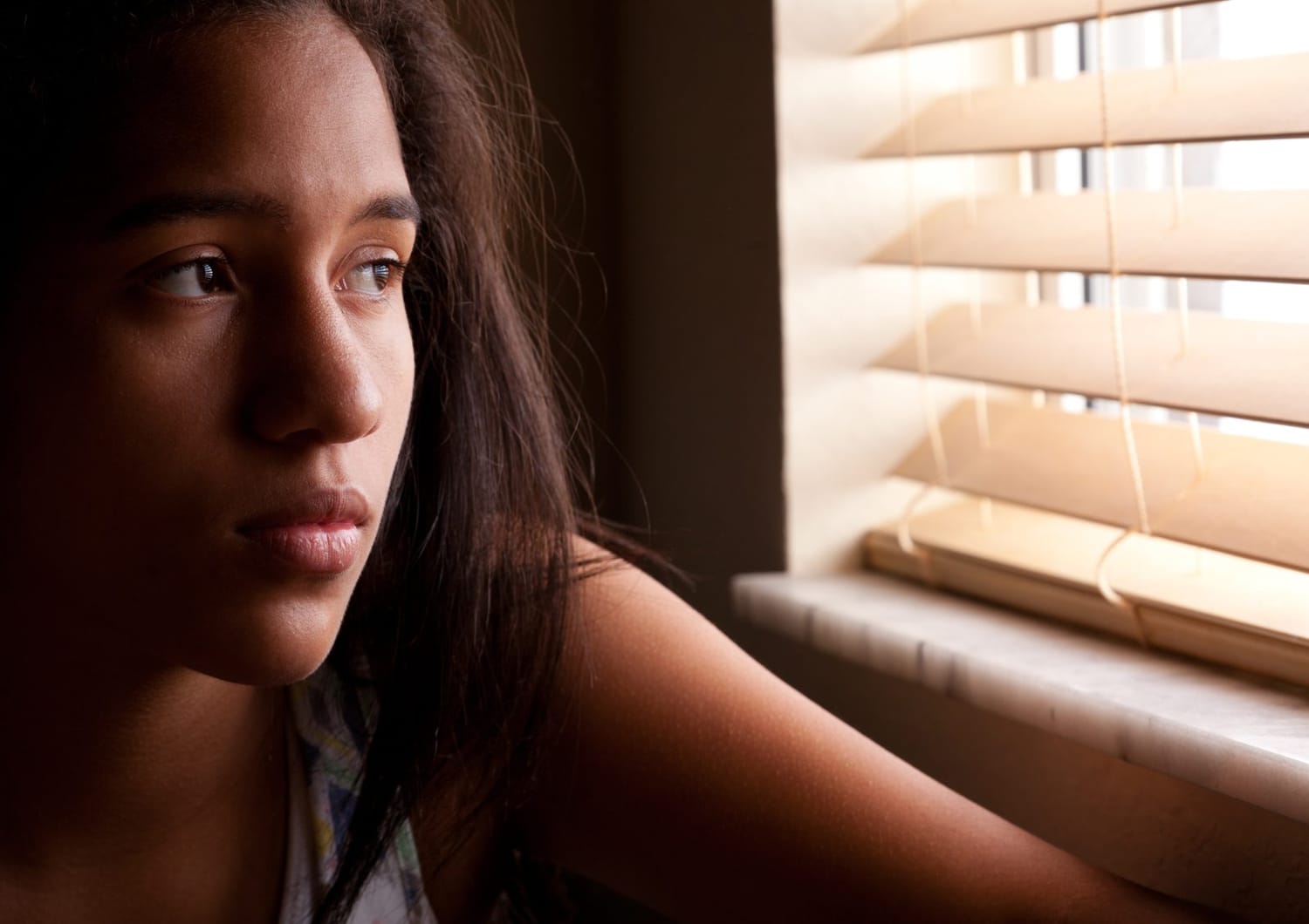 Une adolescente au regard absent, assise près d'une fenêtre à travers les stores