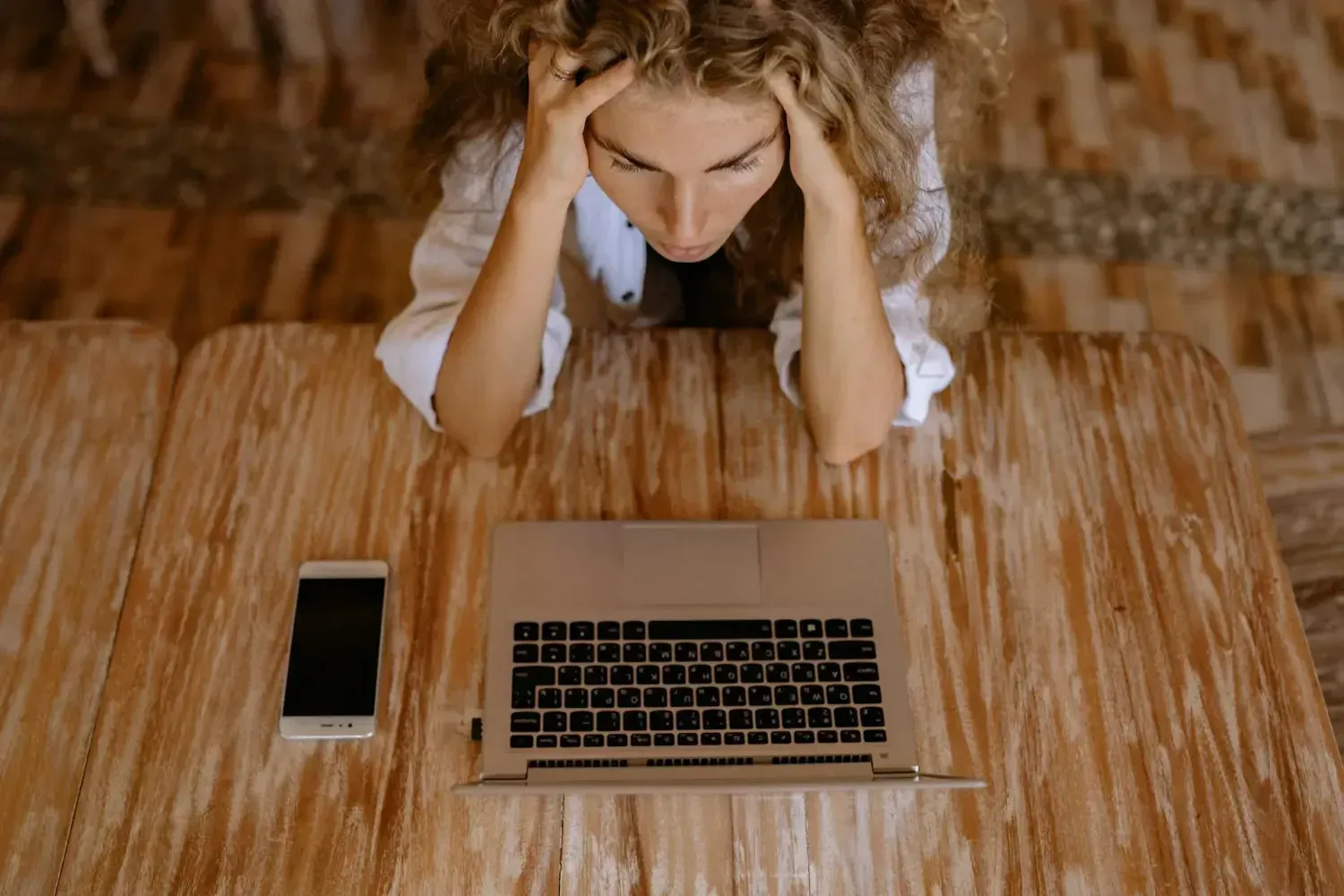 Woman sitting at a wooden desk with her head in her hands, looking overwhelmed in front of a laptop