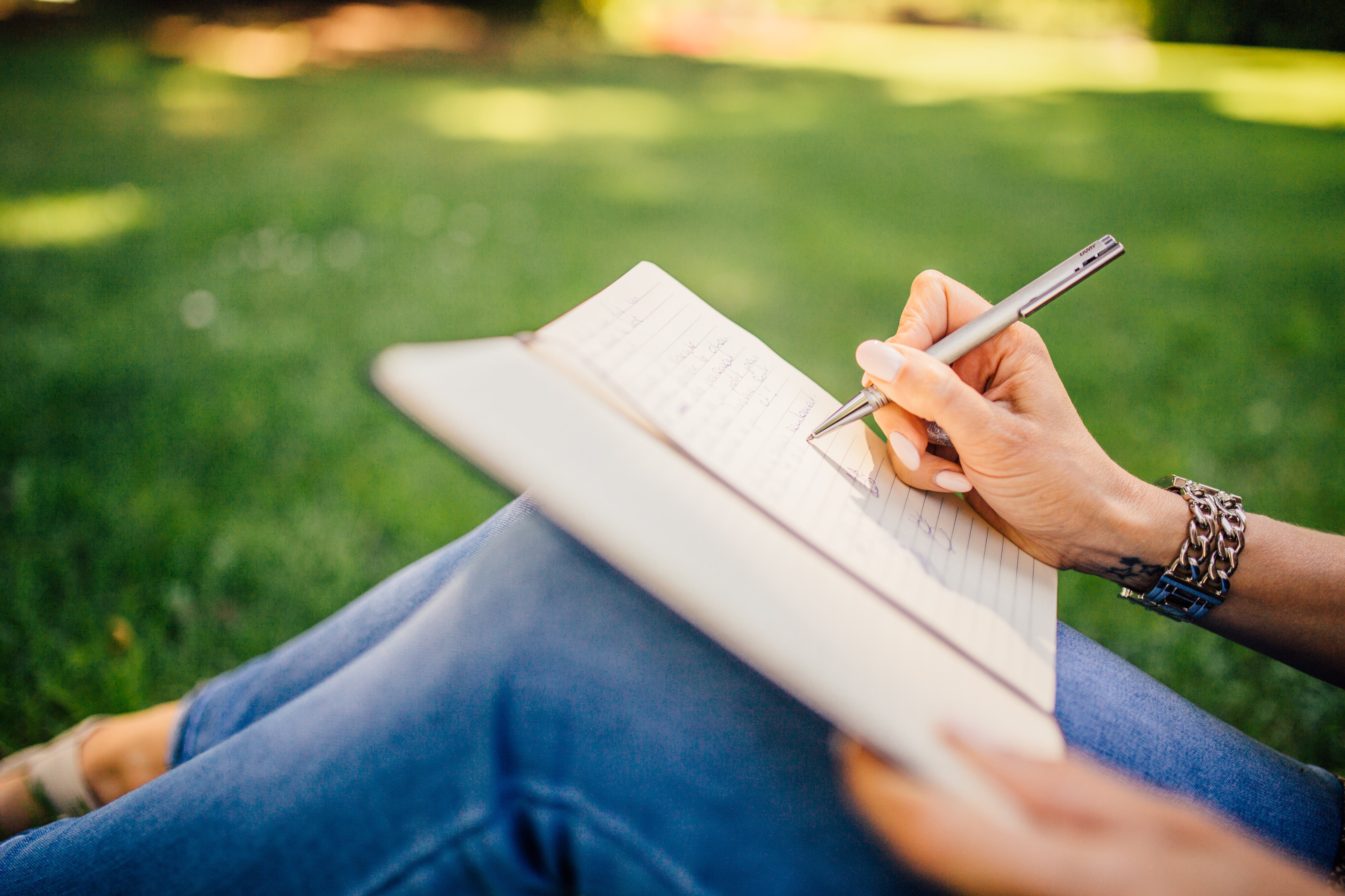 A woman sitting in a park, writing in her notebook. She is engaged in journaling, capturing her thoughts and experiences.