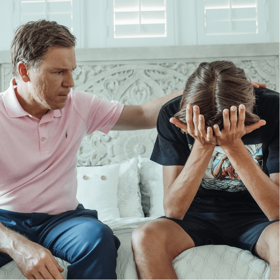 Padre e figlio seduti sul letto. Il padre consola il figlio con una mano sulla schiena mentre il ragazzo preoccupato si sorregge la testa con le mani.