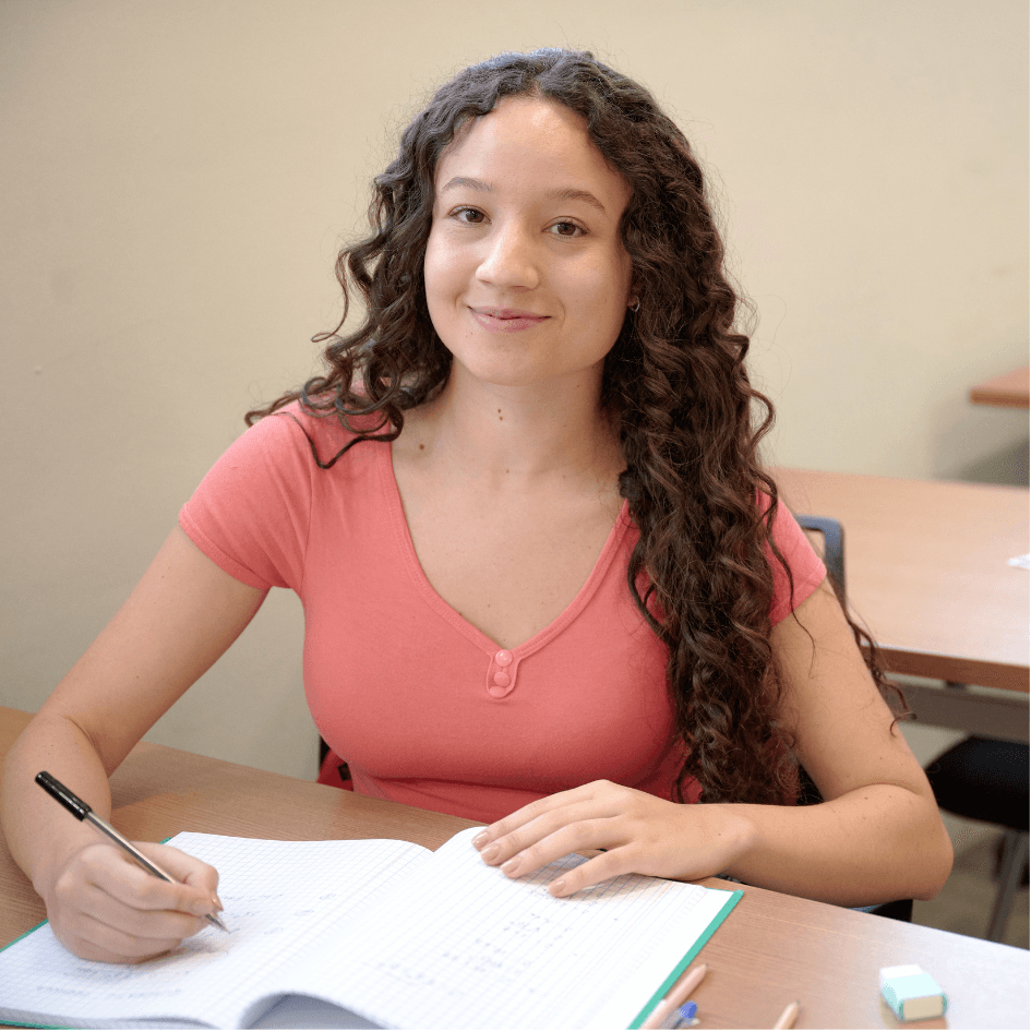 Ragazza con un lieve sorriso e seduta al banco di scuola. Sta svolgendo una verifica in modo sereno e tranquillo.