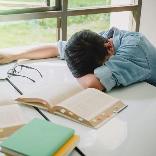 Ragazzo stanco con la testa sul banco mentre studia, circondato da libri aperti e occhiali, in difficoltà nello studio.