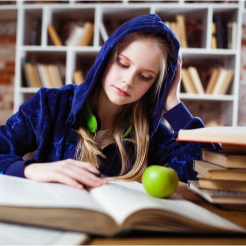 Ragazza concentrata mentre studia, con felpa, circondata da libri e una mela verde sul tavolo.