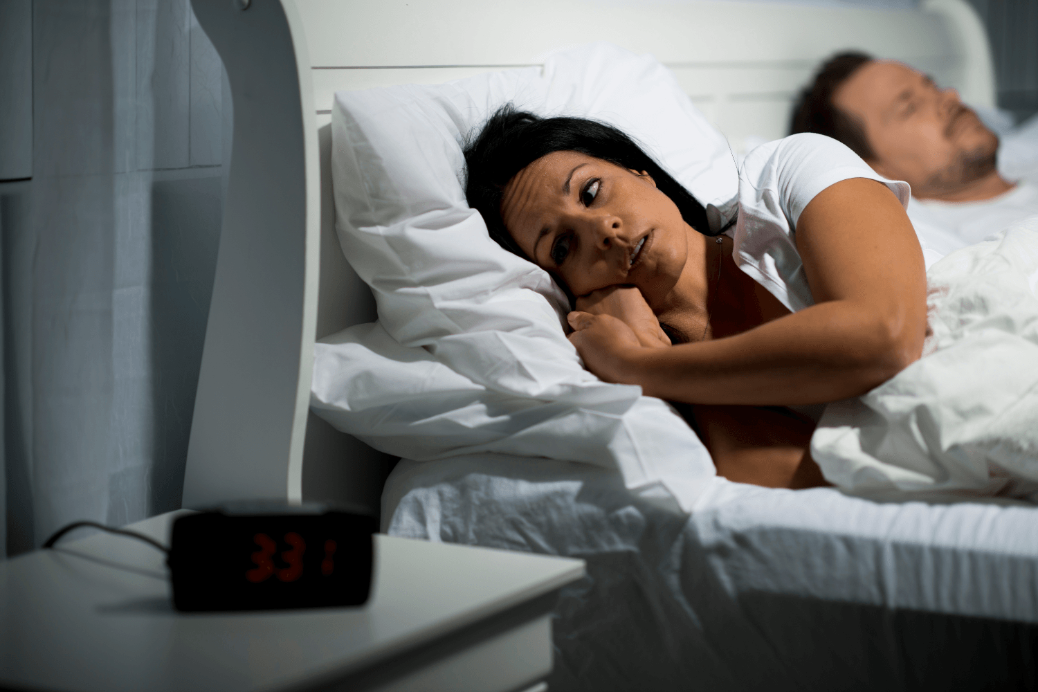 A woman lies awake in bed, looking thoughtful, next to a sleeping man. The clock beside them shows 3:31 AM, suggesting insomnia or restlessness.