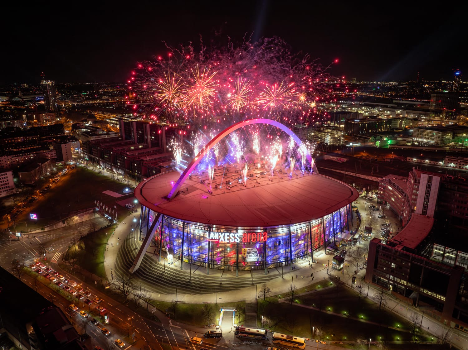 Drohnenaufnahme der LANXESS Arena Köln, während Feuerwerk über dessen Dach explodiert.