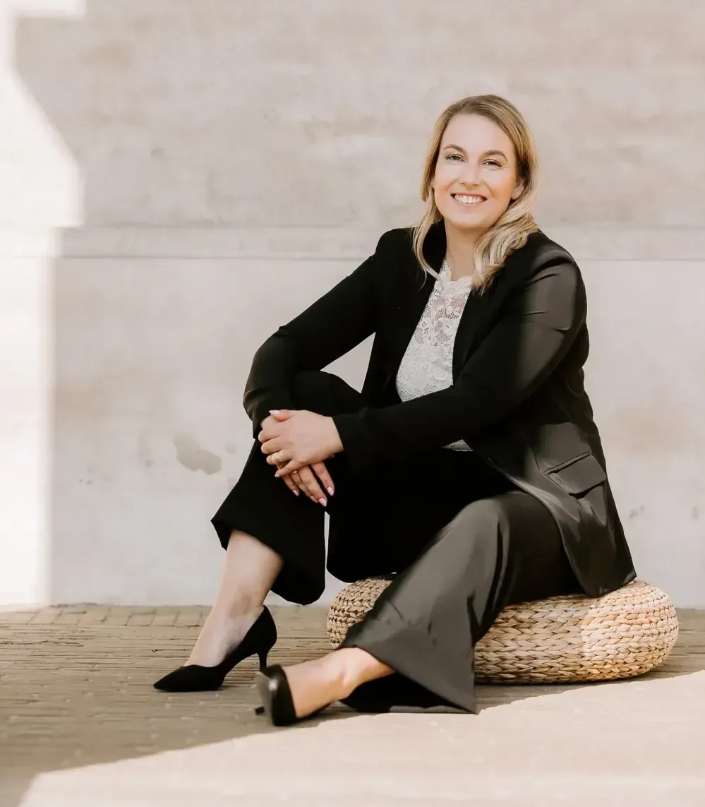 Carina står afslappet foran en lys, hvidmalet murstensvæg. Hun har langt, blondt hår, som er redt tilbage bag skuldrene. Hun smiler venligt mod kameraet. Hun er iført en sort, kortærmet bluse med V-udskæring og sorte bukser. Hun står med vægten let på det ene ben og den ene hånd i lommen. Hun skal sammen med Sandra, Din IT & SoMe Buddy, holde 3 workshops på Nyvej 11A, st., 4450 Jyderup i efterår og vinter 2025 om brugen af Facebook og Instagram, Reels, Stories, synlighed, tekster, Canva og indholdsproduktion hos – Din IT & SoMe Buddy