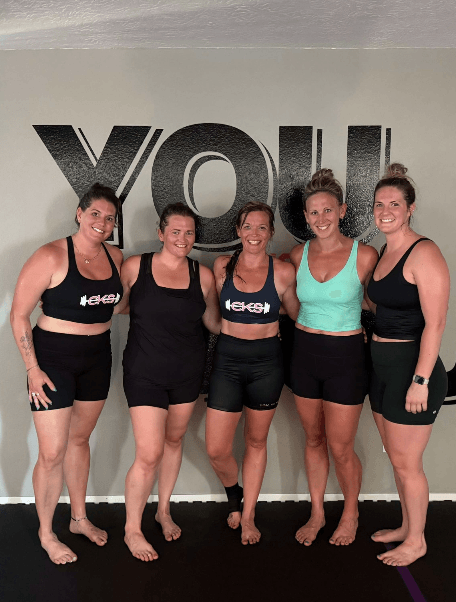 Five women in athletic wear posing together after class in front of the “YOU vs. YOU” wall