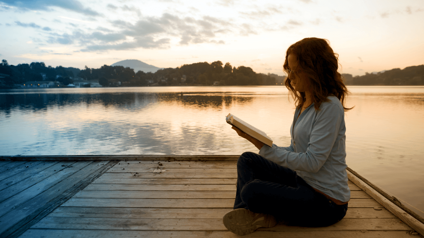  A woman sitting cross legged on a dock looking at a book in her hands, with peaceful water beyond her, followed by mountains and then sky in the distance.