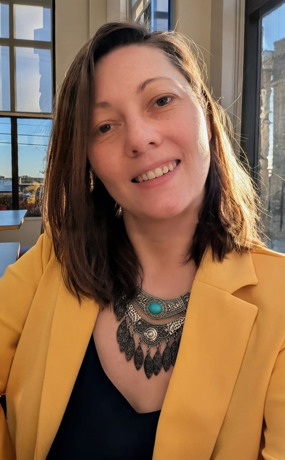 Anna Bohuslavska, inner guide for conscious moms, smiling in a yellow blazer and silver necklace with a turquoise stone, in a sunlit room