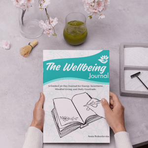Cover of The Wellbeing Journal by Anna Bohuslavska, held in two hands on a table with cherry blossoms and a cup of matcha