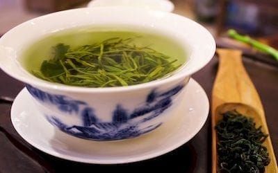 Egy kínai csészében, gyulladáscsökkentő zöld tea van benne van még a teafű is. Mellette egy fakanálban zöld teafű.