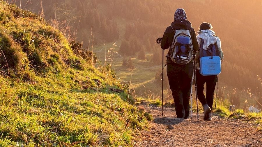 Két hátizsákos túrázó napfényben sétál a hegygerincen; aranyló, derűs táj és tág panoráma – jókedvű kirándulás a természetben