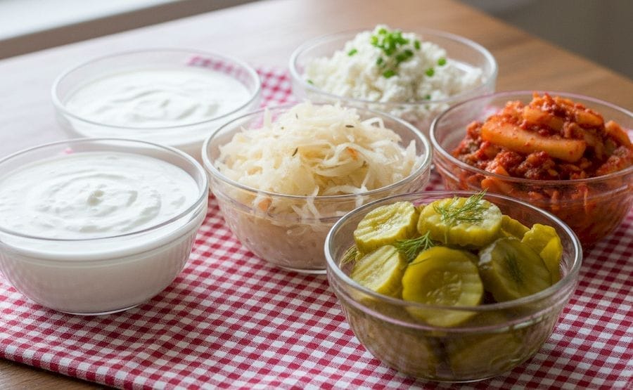 Az asztalon kockás abroszon hat üvegtálkában probiotikumban gazdag, fermentált ételek, savanyúkáposzta, kimcsi, kovászos uborka, kefír, joghurt, található.