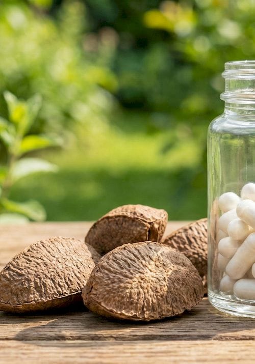 Üvegben lévő fehér szelén kapszulák és egész, illetve feltört paradiók egy fa asztalon, napos kerti háttérrel.