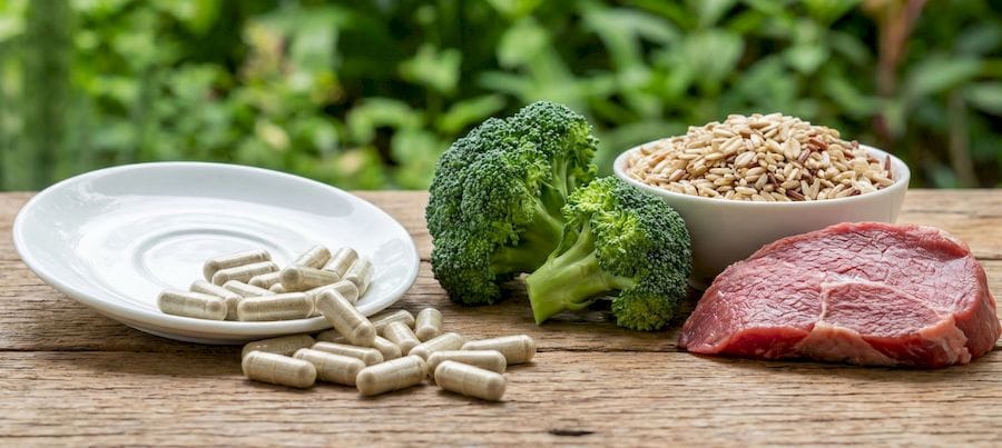 Szerves króm kapszulák egy fehér kistányéron és fa asztalon, mellette természetes forrásai: brokkoli, teljes kiőrlésű gabonák tálban és nyers marhahús szelet.