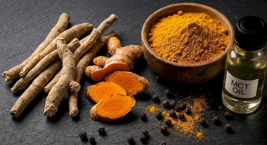 Makrófotó nyers ashwagandha gyökerekről, élénk narancssárga kurkuma szeletekről, aranyló kurkumaporról, fekete borsról és MCT olajról egy sötét pala felületen.