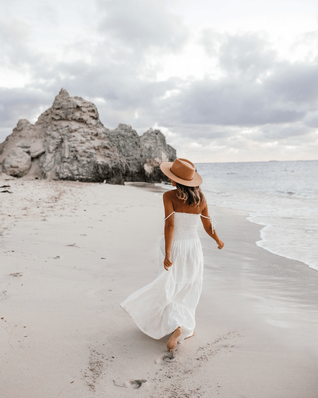 Femme en robe blanche marchant pieds nus sur une plage au lever du jour.