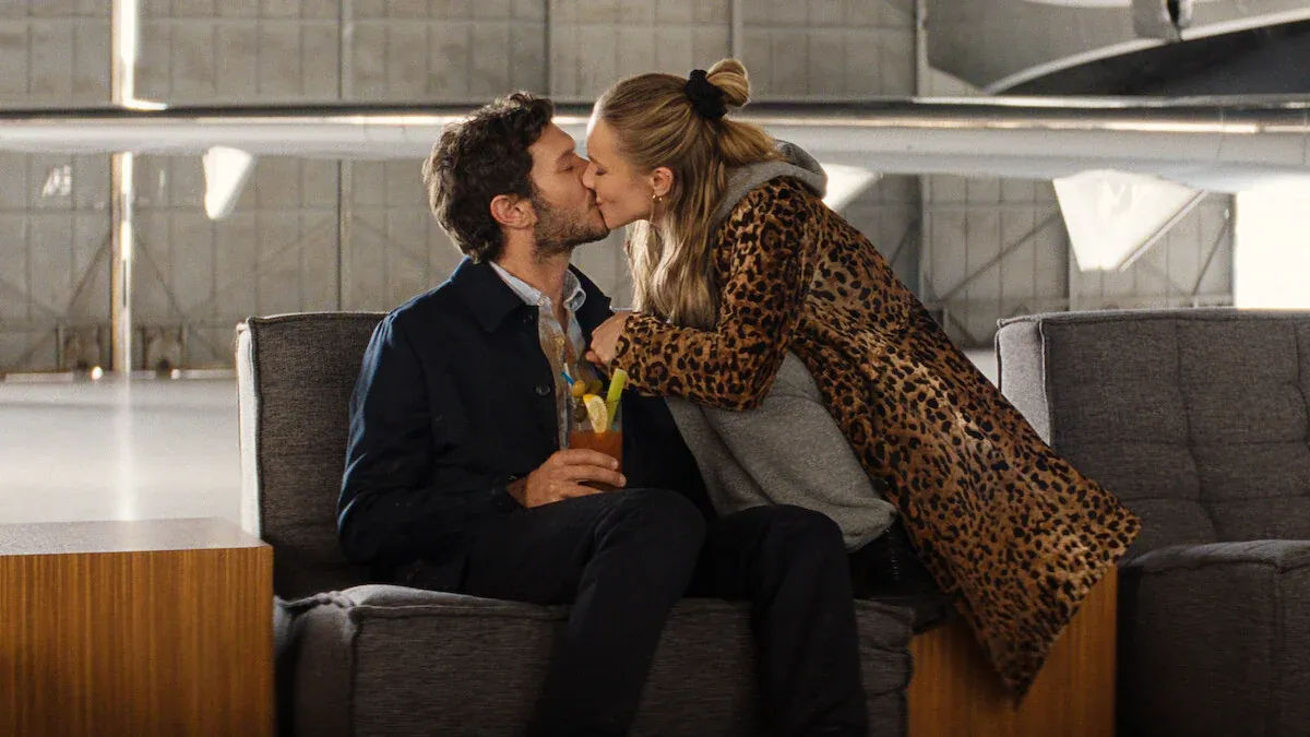 Joanne, con un cappotto leopardato, bacia Noah seduto con un Bloody Mary in mano in un hangar dell'aeroporto, in Nobody Wants This 3