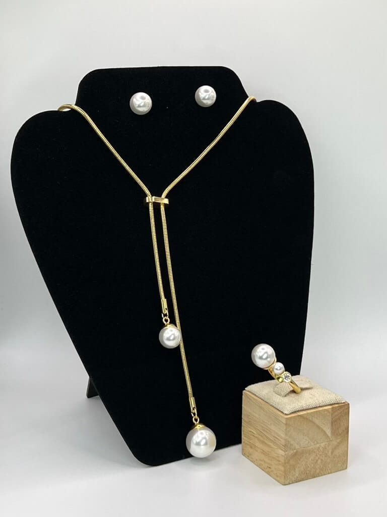Parure de bijoux en acier inoxydable plaqué or avec perles blanches : collier, boucles d'oreilles et bague sur fond neutre