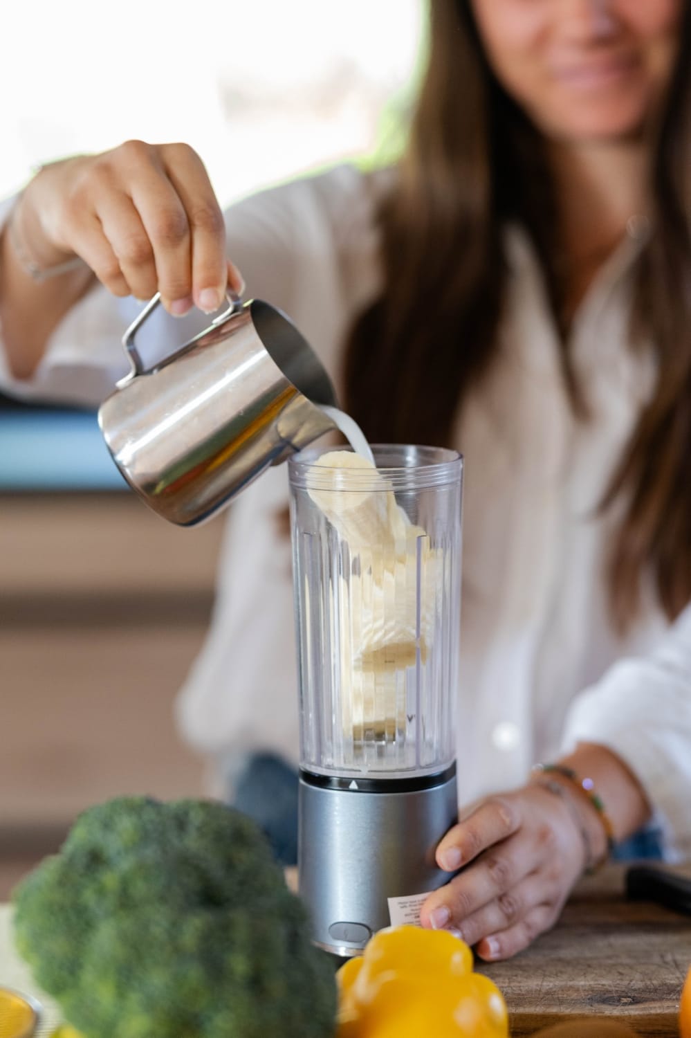 Mélanie Padovani verse du lait végétal dans un blender pour préparer un smoothie, un brocoli au premier plan